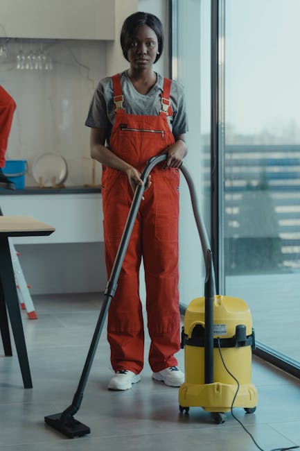 A professional cleaner from Newbury Park Carpet Cleaners is vacuuming a modern, wooden floor in a bright kitchen area with large windows. The cleaner is wearing a grey t-shirt and red overalls, holdin