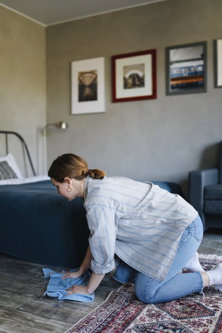 A person kneeling on a patterned carpeted floor, holding a yellow and black portable vacuum cleaner with a hose attachment. The individual is wearing a beige jacket and blue jeans, with their head sli