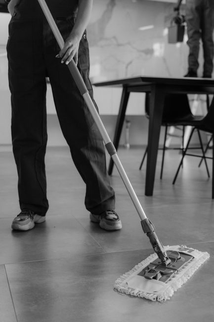 A person dressed in protective clothing, including gloves and disposable jumpsuit, is using a handheld vacuum cleaner to perform surface cleaning in a modern indoor setting. The vacuum has a long hose
